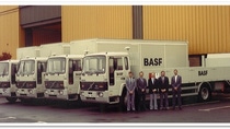 Historic photograph of five BASF-branded lorries parked outside an industrial building, with five people standing in a row in front of the central vehicles. 