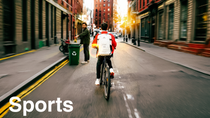 A cyclist with a red jacket and white backpack, marked with a glowing light-ring effect, rides down a city street at sunset, past a person recycling in the background. The text reads: "Sports."