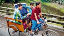 Ein Mann fährt Fahrrad während zwei weitere Männer in einem Anhänger sitzen und eine Solarplatte halten Ein Mann fährt Fahrrad während zwei weitere Männer in einem Anhänger sitzen und eine Solarplatte halten