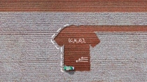 The picture is an aerial view of a cotton field, where a harvesting machine is working. The soil showing where the cotton has been harvested is in the shape of a T-shirt. There are white handwritten sketches on the picture showing scientific formulae and three arrows. 