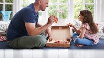 Father and daughter sitting on a sofa eating pizza.