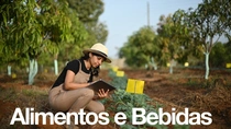 Uma agricultora, vestindo macacão e chapéu, segura um tablet e examina plantas em um campo em um dia ensolarado. O texto na imagem diz: “Alimentos e bebidas”.