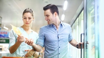 Early 30's couple buying frozen food in local supermarket.They are standing next to freezer section and reading labels and price tags, while she's still uncertain of which product to pick. Both casually dressed Early 30's couple buying frozen food in local supermarket.They are standing next to freezer section and reading labels and price tags, while she's still uncertain of which product to pick. Both casually dressed