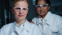 Two female scientists wearing white lab coats with the BASF logo and safety goggles stand in the laboratory and look into the camera.