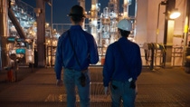 Two men with satety helmets are walking side by side in a lit production facility Two men with satety helmets are walking side by side in a lit production facility