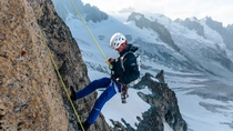 Climber ascending a steep rock face in an alpine mountain landscape, secured with rope and equipment. 