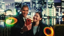 A man and a woman in casual jackets stand in front of an Asian city skyline at night. They are both looking at his phone. Light swirls appear next to them.