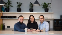 Three employees stand together at a counter in a modern office kitchen, smiling and engaging in a casual conversation, with plants and shelves in the background.