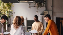 En la foto un grupo de cuatro personas mirando la computadora portátil. De izquierda a derecha, un joven negro de camisa roja, una mujer negra de pelo rizado, una mujer blanca de pelo castaño y blazer gris y poco después un hombre blanco de mediana edad, pelo y barba blanca y chaqueta negra. 