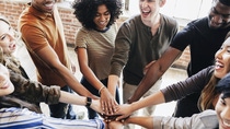 A diverse group of young people stands in a circle, placing their hands on one another’s as a sign of teamwork and solidarity, laughing and smiling.