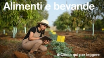 Una agricoltrice, con indosso una salopette e un cappello, tiene in mano un tablet mentre osserva le piante in un campo in una giornata di sole. Il testo sull’immagine recita: “Cibi e bevande”.  Una agricoltrice, con indosso una salopette e un cappello, tiene in mano un tablet mentre osserva le piante in un campo in una giornata di sole. Il testo sull’immagine recita: “Cibi e bevande”.
