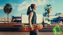 A young woman with headphones walks along a city street holding a skateboard. In the background there are palm trees, buildings, and a truck. A light swirl appears on the road surface.