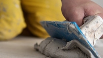 A person kneels on the floor wearing yellow pants, using a blue trowel to spread grey cement. The focus is on the hands and tools.