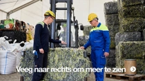 Twee mannen in gele helmen en werkoveralls staan naast een groot, geperst blok textielafval.  Twee mannen in gele helmen en werkoveralls staan naast een groot, geperst blok textielafval.