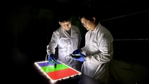 Zwei Wissenschaftler in weißen Laborkitteln und blauen Handschuhen untersuchen in einem abgedunkelten Raum eine leuchtende Farbfläche in Rot, Grün und Violett. Einer hält ein Messgerät. Zwei Wissenschaftler in weißen Laborkitteln und blauen Handschuhen untersuchen in einem abgedunkelten Raum eine leuchtende Farbfläche in Rot, Grün und Violett. Einer hält ein Messgerät.