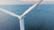Close-up of the rotor of a wind turbine taken from bird perspective, with more wind turbines in the backdrop, standing in a row in a sea with waves. 