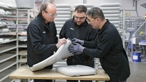 Drei Männer in schwarzen Vitra-Uniformen und Schutzbrille untersuchen gemeinsam ein weißes Stuhlschalenmodell in einer Produktionshalle. Im Hintergrund sind Regale mit weiteren Bauteilen zu sehen. Drei Männer in schwarzen Vitra-Uniformen und Schutzbrille untersuchen gemeinsam ein weißes Stuhlschalenmodell in einer Produktionshalle. Im Hintergrund sind Regale mit weiteren Bauteilen zu sehen.