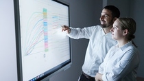 A man and a woman are discussing data charts on a large monitor.