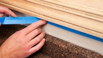 A painter is applying blue masking tape to protect an area of wall below oak window trim which is being refinished. The wall repainted after staining the trim, below the hand is a granite countertop.