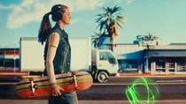 A young woman with headphones walks along a city street holding a skateboard. In the background there are palm trees, buildings, and a truck. A light swirl appears on the road surface.