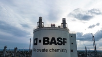 Wide view of an industrial BASF facility with a large storage tank labeled “BASF – We create chemistry,” where several workers in safety helmets stand together on the top platform under a cloudy sky.