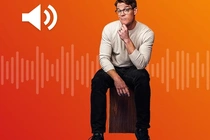 Ein Mann mit Brille sitzt auf einer Holzkiste vor einem orangefarbenen Hintergrund. Links oben ist ein weißes Lautsprecher-Icon zu sehen, dahinter eine Audiowellenvisualisierung. Ein Mann mit Brille sitzt auf einer Holzkiste vor einem orangefarbenen Hintergrund. Links oben ist ein weißes Lautsprecher-Icon zu sehen, dahinter eine Audiowellenvisualisierung.