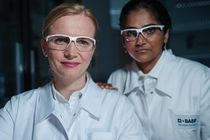Two female scientists in white lab coats with the BASF logo and protective goggles stand in the laboratory and look into the camera.
