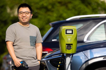 A man with an electric charger in his hand stands in front of electric cars at an electric charging station.