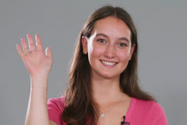 A young woman stands in front of a neutral background and smiles at the camera. She has raised her hand in a friendly greeting.