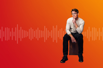 A man sits on a wooden stool in front of a red-to-orange gradient background with stylized sound waves stretching horizontally across the image. A man sits on a wooden stool in front of a red-to-orange gradient background with stylized sound waves stretching horizontally across the image.