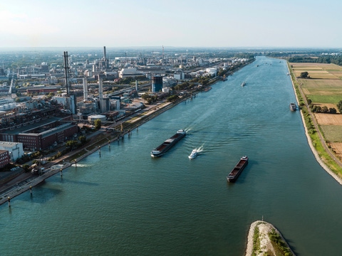 Aerial view of the BASF chemical production site in Ludwigshafen along a river, with cargo ships on the water and industrial facilities extending across the landscape.