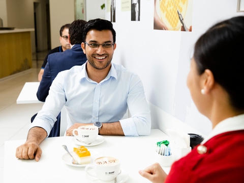 Two people sit across from each other in a bright cafeteria, smiling and talking over coffee and cake, with other people seated in the background.