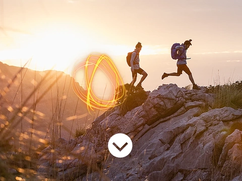 Two people wearing VAUDE backpacks run across rocky terrain in a mountain landscape at sunset. An orange light swirl is visible in the foreground.