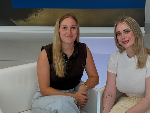 Alena Wolf und Tamara Rettig sitzen auf weißen Sesseln in modernem Raum mit großem Wandbild im Hintergrund