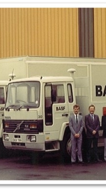 Photograph from 1986 showing five BASF delivery lorries parked outside an industrial warehouse, with five employees standing in front of the vehicles. BASF branding is visible on the trucks and warehouse loading area.