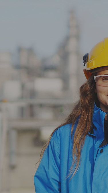 An engineer wearing a hard hat and safety goggles stands in front of an industrial plant.