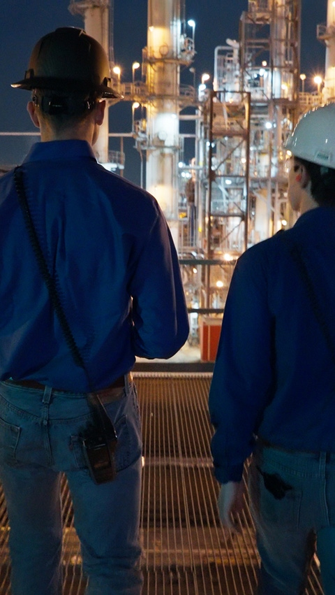 Two men with satety helmets are walking side by side in a lit production facility