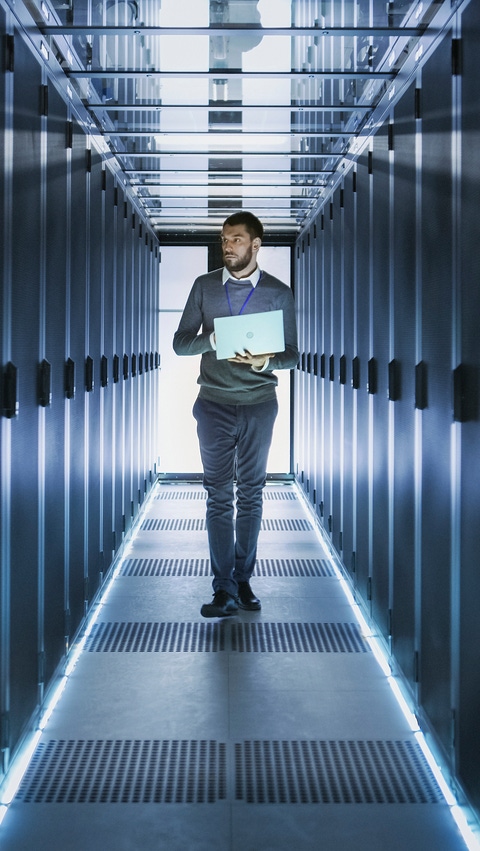 A technician carrying a laptop walks through a modern data center surrounded by illuminated server racks. A technician carrying a laptop walks through a modern data center surrounded by illuminated server racks.