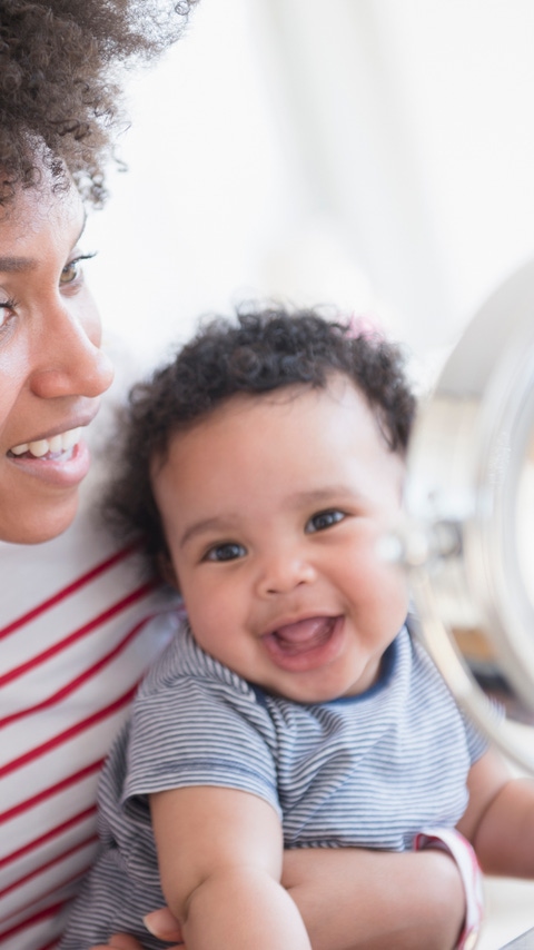 Mother holding baby son applying face cream in mirror