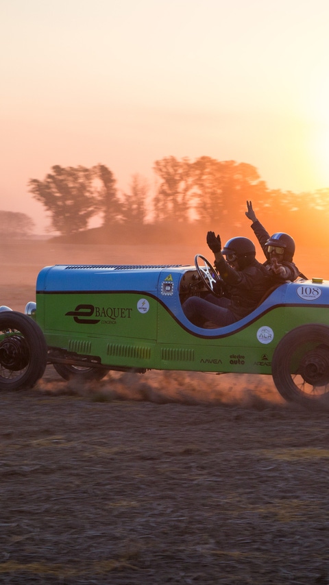 Partly silhouetted against the sunset, racing driver Hernan Bradas and his co-driver wave as they drive the green and blue eBaquet, an electric car built in the style of a famous 1940s racing car, along a dusty track in Argentina.
