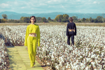 Two models wearing yellow and black clothes made from cotton are walking along a catwalk in a field of fluffy white cotton plants with blue mountains in the background. 