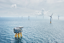 A wide view of a blue ocean with waves taken from above, showing two rows of wind turbines stretching into the distance and an offshore platform with helicopter landing pad in the foreground. A wide view of a blue ocean with waves taken from above, showing two rows of wind turbines stretching into the distance and an offshore platform with helicopter landing pad in the foreground.
