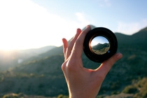 A hand is holding a lens in front of a mountain, focusing on the summit