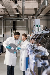 Superconductors transmit electric power with negligible line losses and have high current-carrying capacity compared to copper. This will make it possible to generate and transmit electricity in a resource-saving manner in future.
For the efficient production of high-temperature superconductors Deutsche Nanoschicht GmbH, a BASF Group company, has developed a unique process: very thin films of a superconducting material are applied to a metal strip in a continuous process by a technique called chemical solution deposition. In contrast to other, physical methods, the chemical process requires neither a vacuum nor a clean room environment. This provides a decisive advantage for the production costs of the superconducting wires.
Anushka Kudaliyange (left) and Margot Ledwon-Brzezina (right), both employees of Analytics and Pilot Production, examine metal substrates before coating.
