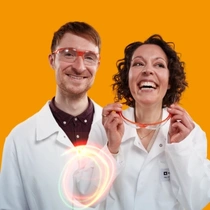 Ein männlicher und eine weibliche BASF-Mitarbeitende aus Deutschland stehen in weißen Laborkitteln vor einem orangefarbenen Hintergrund. Ein Lichtwirbel schwebt um ihre Körper.