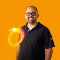 Um colaborador da BASF sorri olhando para a câmera, vestindo uma camisa polo azul-marinho com o logo da BASF na manga, em frente a um fundo laranja. Um redemoinho de luz em tons de vermelho, laranja e verde aparece ao seu lado.