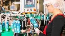 A female production worker wearing protective gloves, safety glasses and hairnet inspects a bottle of green-blue colored shower gel on a production line. A female production worker wearing protective gloves, safety glasses and hairnet inspects a bottle of green-blue colored shower gel on a production line.